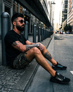 Street portrait of a man in sunglasses seated on a city pavement, edited with Dark Tones Lightroom presets