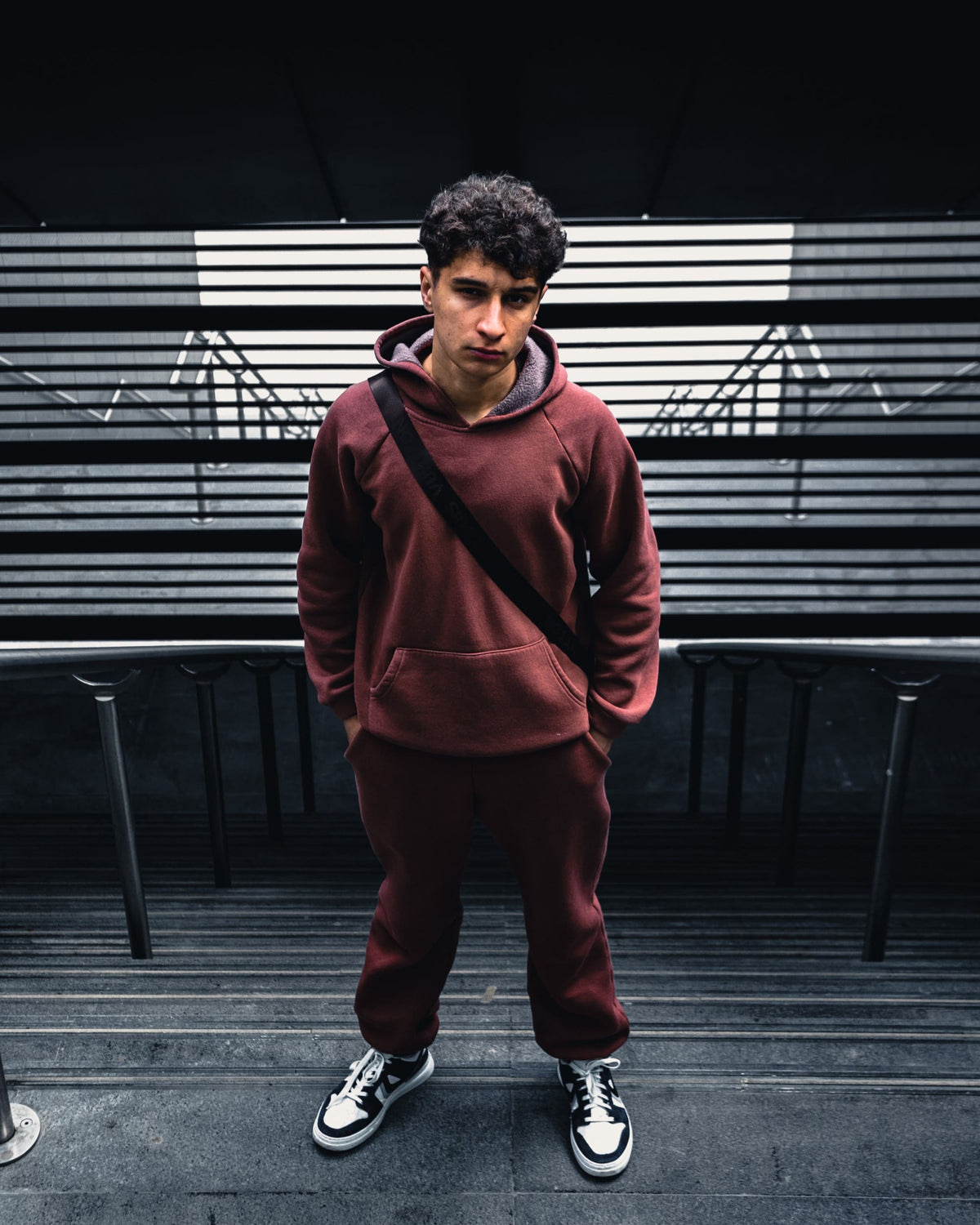Street portrait of a man in a burgundy tracksuit at the top of a flight of stairs, with deep shadows and subdued colours