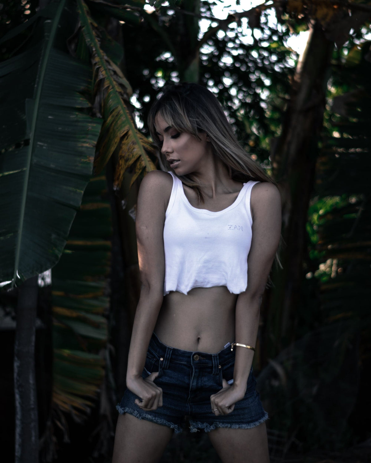 Moody portrait of a woman in a white crop top among dark, desaturated tropical leaves