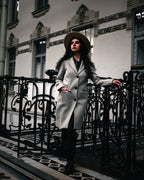 Woman in a light coat and hat beside iron railings, with muted architectural details and deep shadows