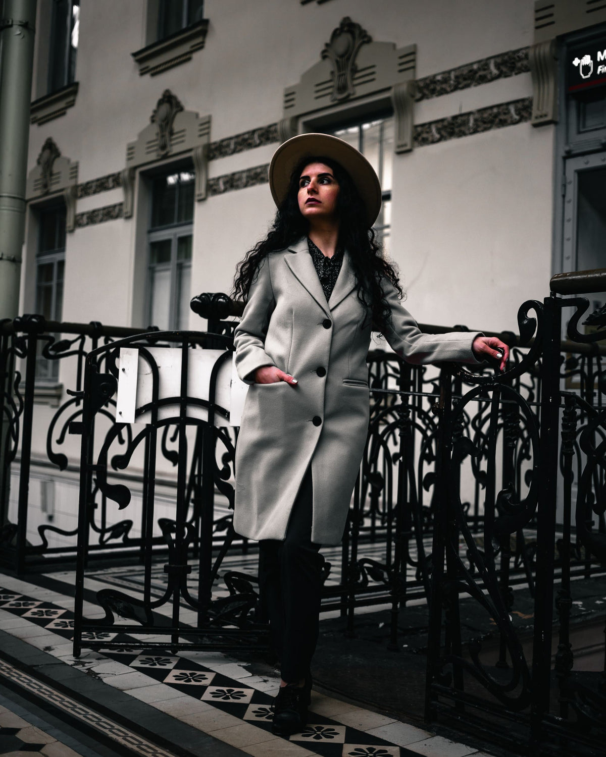 Woman in a light coat and hat beside iron railings, with muted architectural details and deep shadows