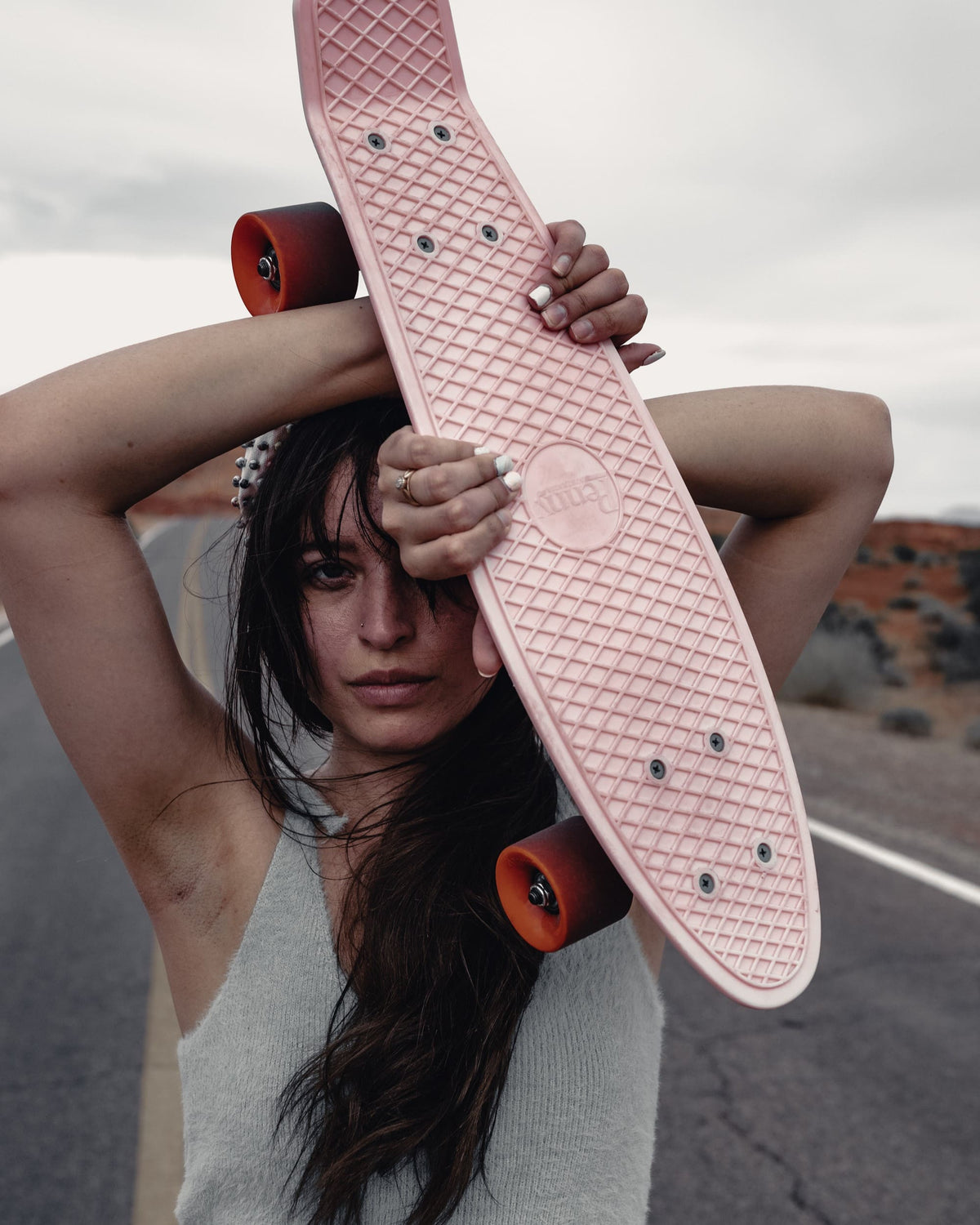 Moody portrait of a woman holding a pink skateboard above her head, with subdued colours and deep shadows