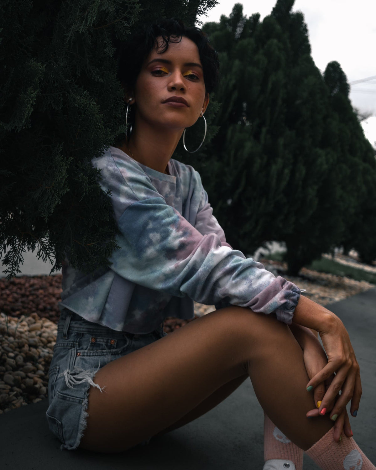 Moody outdoor portrait of a woman in a pastel sweatshirt, framed by dark, desaturated foliage